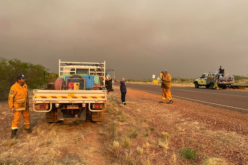 Several firefighters standing around a ute parked on the side of an outback highway, with bushfire smoke in the background.