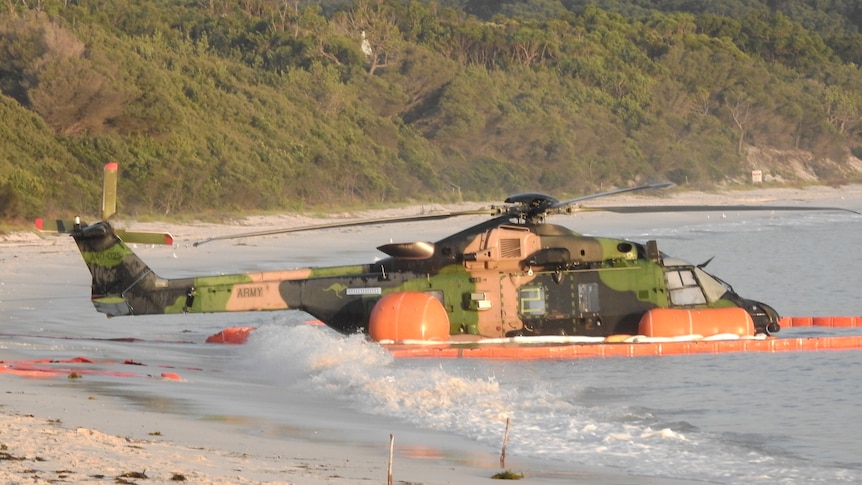 A green and beige camouflaged helicopter sits in shallow water at daybreak at a beach.
