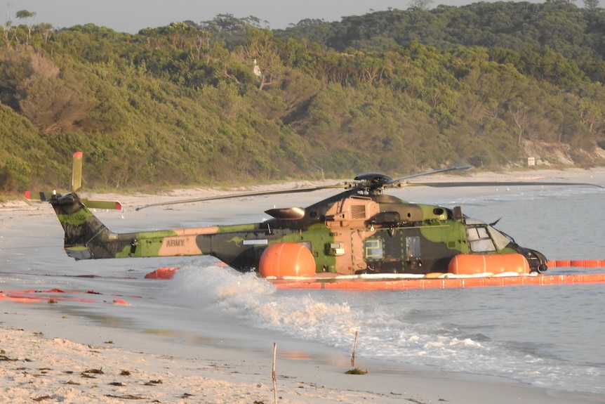 A green and beige camouflaged helicopter sits in shallow water at daybreak at a beach.