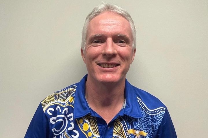 A man with grey hair wears a blue shirt featuring Aboriginal art, while standing in front of a white wall.
