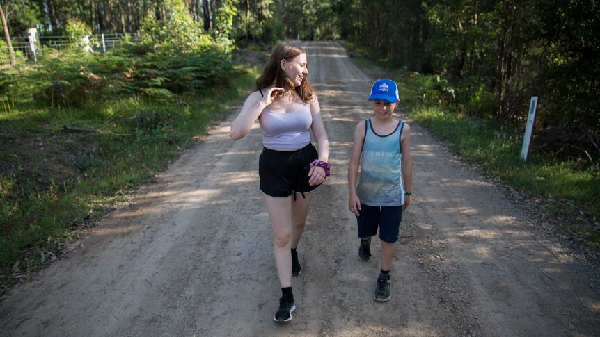 A teenage girl and younger boy are caught in a slice of sunlight as they walk towards the camera along a bush-lined dirt road.