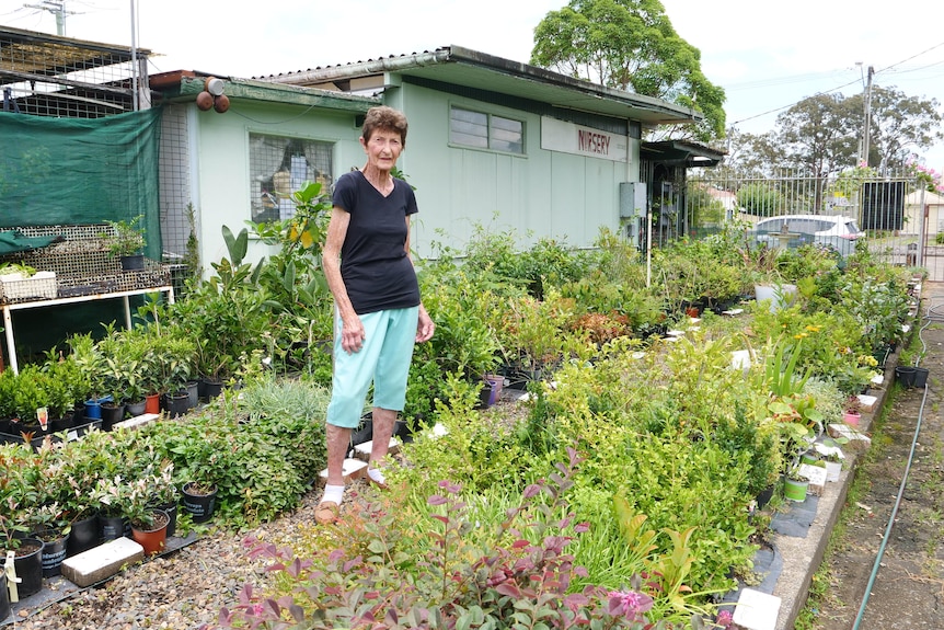 An older woman stands in front of a nursery, between rows of plants.