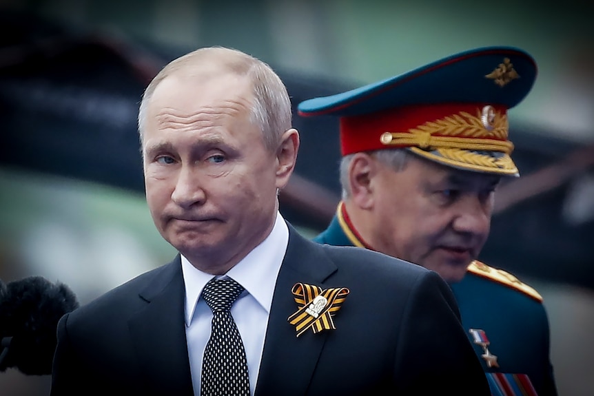 Vladimir Putin looking thoughtful while a Russian military general in dress uniform passes behind him