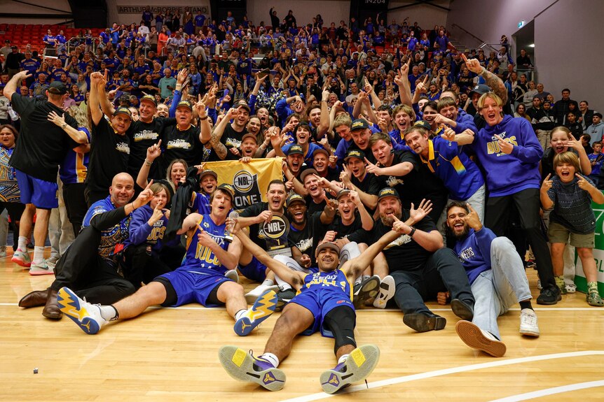A men's basketball team in blue uniforms with their arms in the air and big smiles. A large crowd is celebrating behind them.