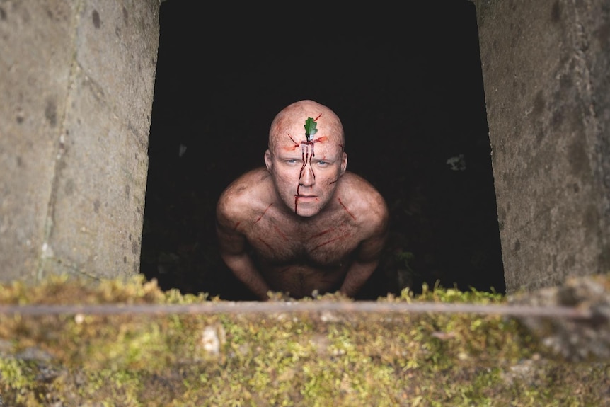 Bald white man stands shirtless and bloodied with a green leaf stuck to his forehead from the inside of a dark pit.