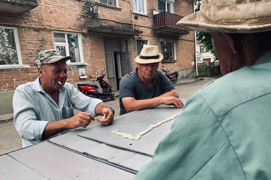 Three men sit at an outside table playing dominos.