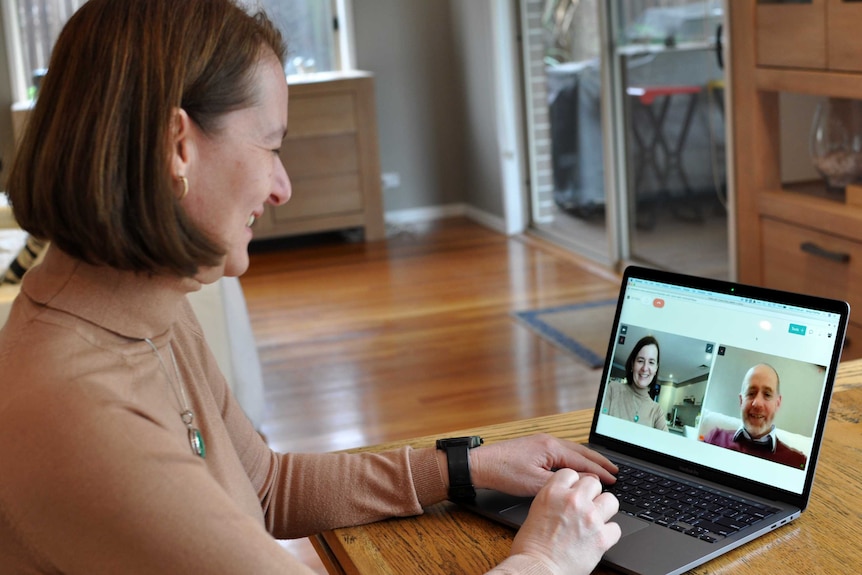 A woman smiles as she looks at a laptop on a table. She is video calling a man through a web browser telehealth program.