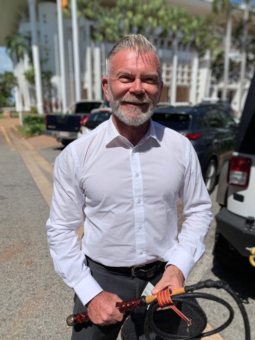 A man in a business shirt smiling and holding a leather whip outside court.