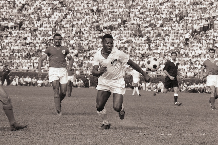 Pele surges forward towards a bouncing ball during a football match, as opposition players watch on in the background.