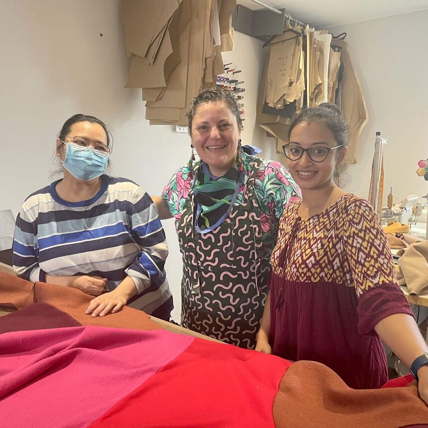 Three women stand smiling in a sewing room in front of colourful material on a table. One wears a facemask.