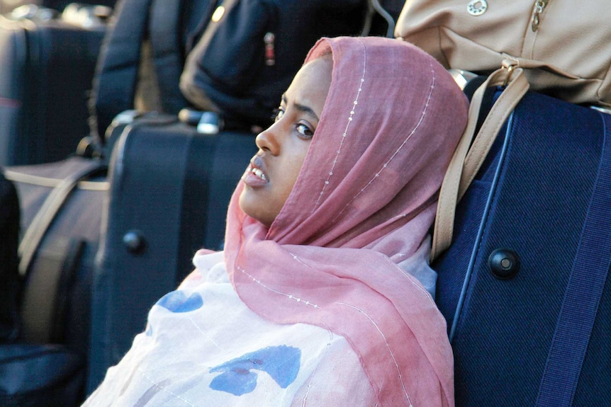 A woman in a hijab leans against a suitcase looking worried