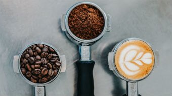 Coffee beans, ground coffee and a latte sit in espresso machine grips on a bench for a story on whether drinking coffee is good