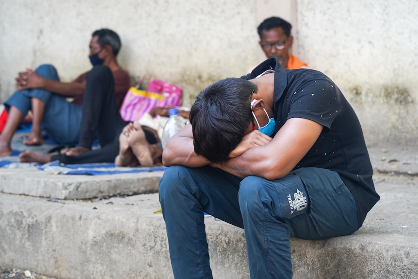 An Indian man in a face mask bends forward to rest his face on his knees