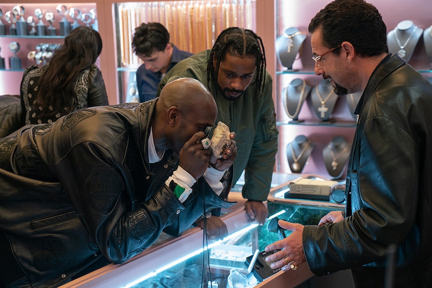 A man in leather jacket bends over and inspects stone with handheld loupe as two men look on in fluorescent lit jewellery shop.