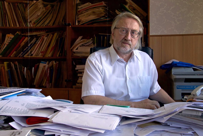 A man with a white beard and glasses wearing a white shirt stares into the camera.