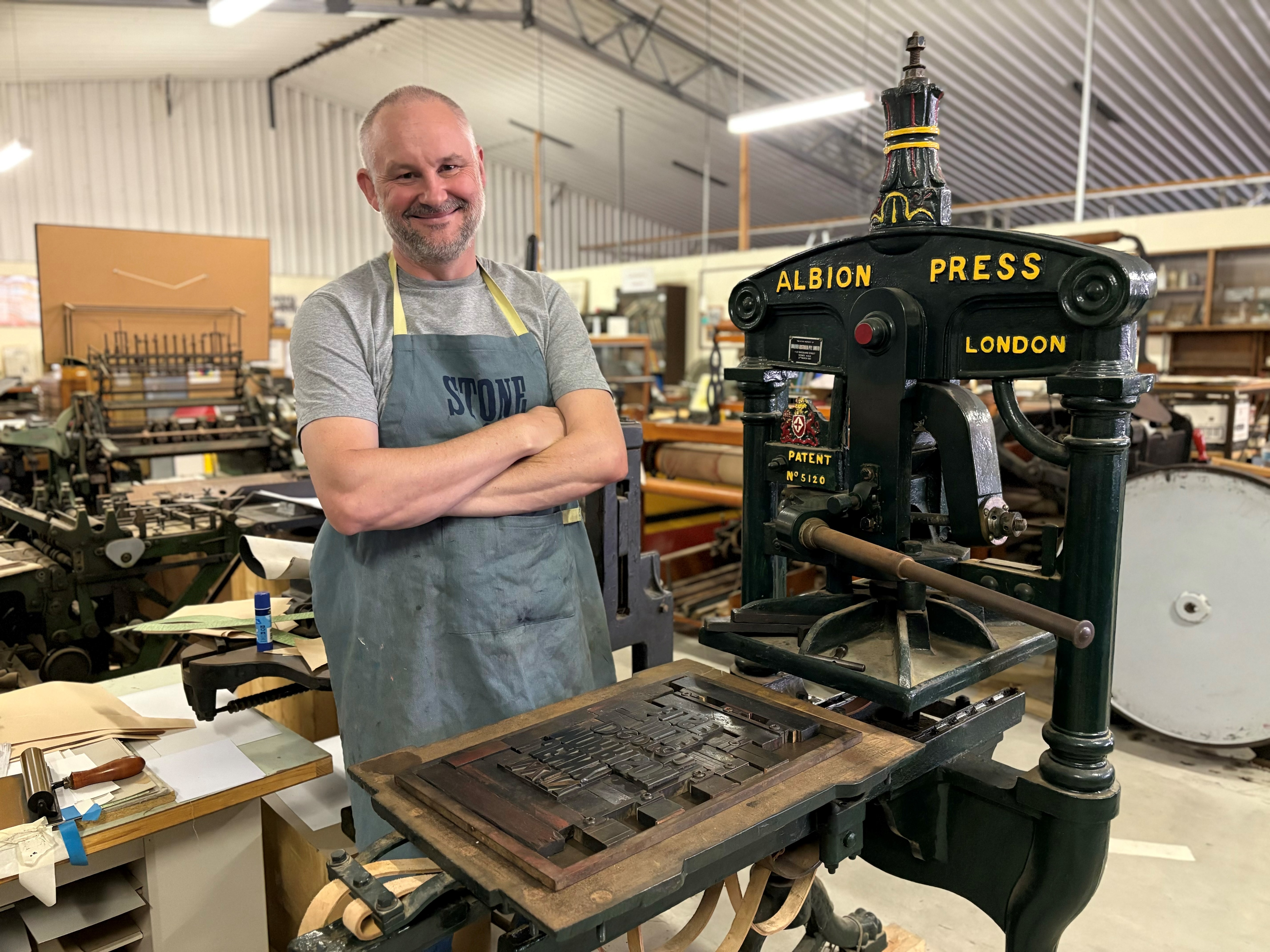 A balding man standing with his arms crossed and an apron on next to an old machine used for print