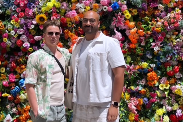 Shane, left, smiles as he stands next to Alex, right, in front of a multicolour flower wall on a sunny day.