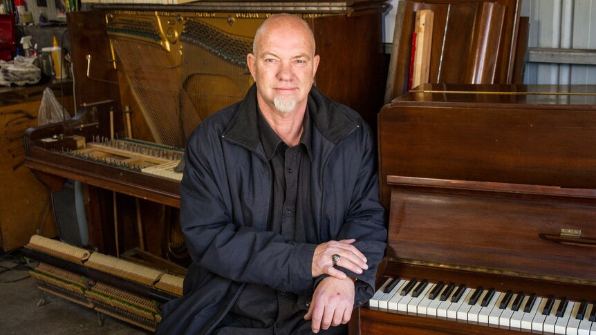 Musician Ray Vanderby stands in front of an old piano which has been taken apart so you can see the strings and inner workings