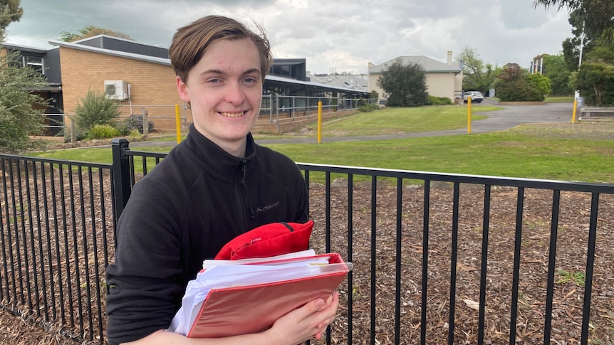 A young man with combed brown hair stands in front of an empty schoolyard. He is holding study books and a pencil case.