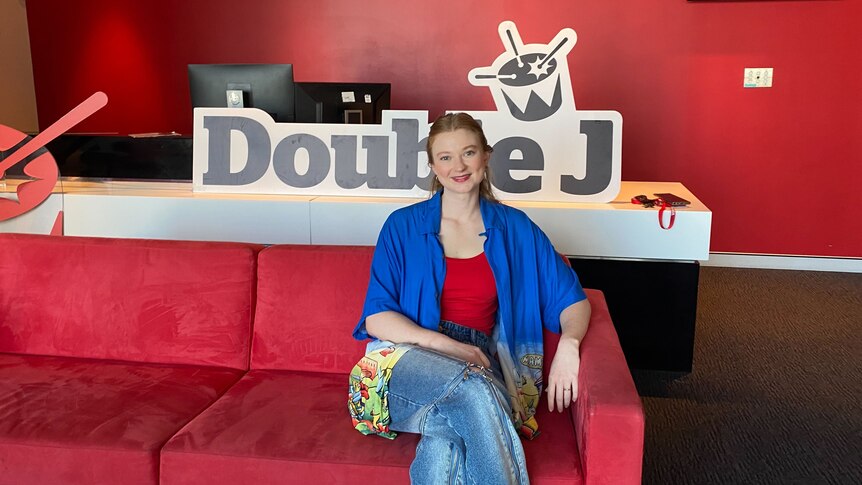 Author Bri Lee sits on the red triple j couch wearing a blue open shirt red singlet and jeans with the Double J sign behind her 