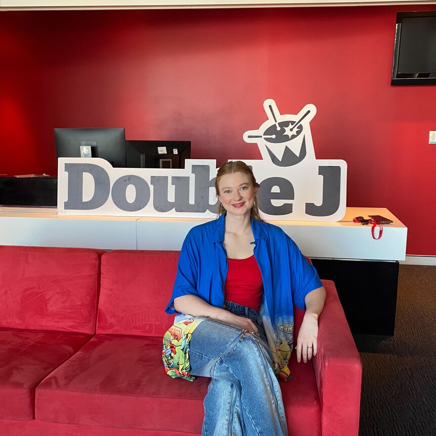 Author Bri Lee sits on the red triple j couch wearing a blue open shirt red singlet and jeans with the Double J sign behind her 