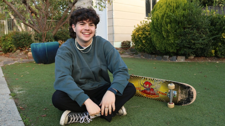 A teenage boy sits next to a skateboard.