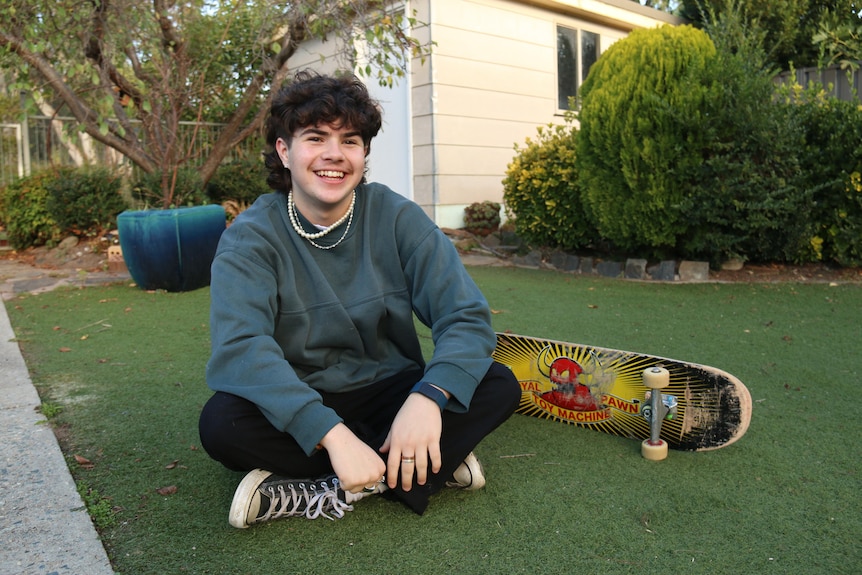A teenage boy sits next to a skateboard.