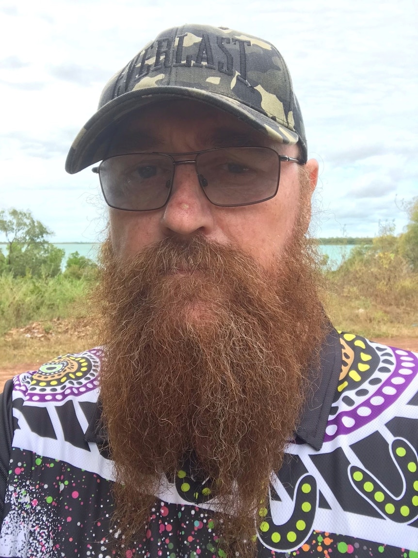 A man with glasses and a beard stands in a rural beach area.