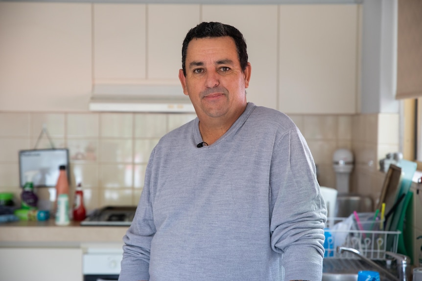 A man in a grey jumper stands in a kitchen.