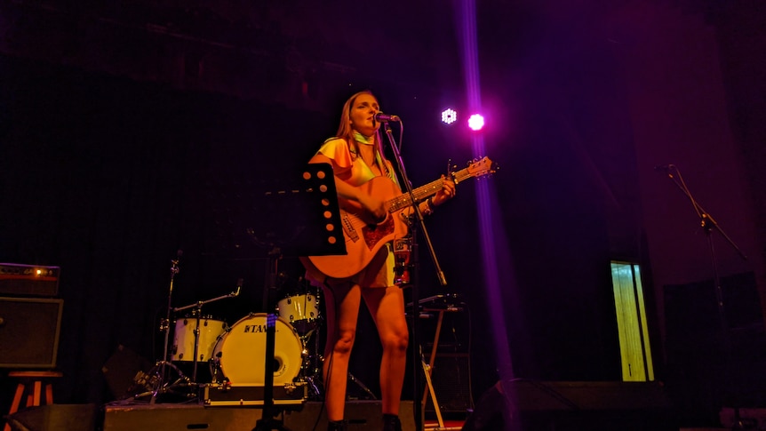 woman stands on a stage singing into a microphone while playing a guitar 