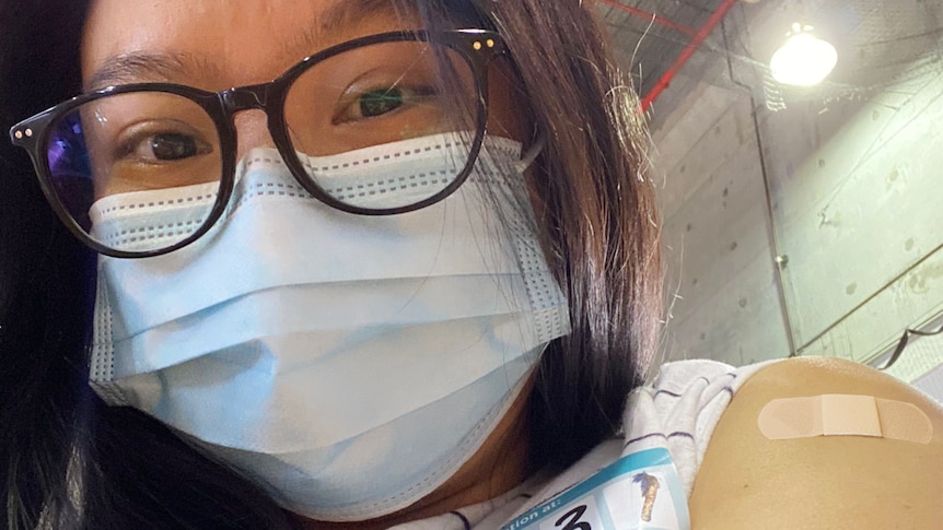 Edwina wearing glasses and face mask showing the bandaid on her arm, in a story about vaccine selfies benefits.