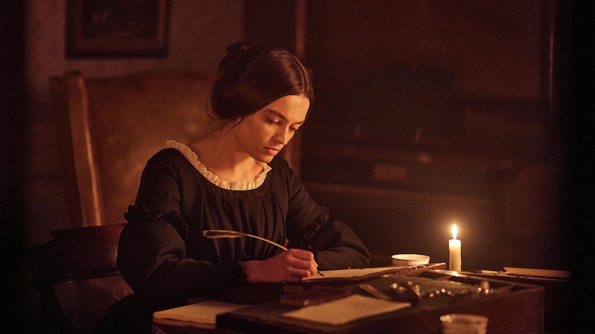 Young white woman with dark hair pulled into  bun wears a black neoclassical 1800s dress and sits writing at a desk with a quill