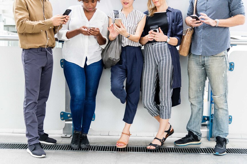 A group of people stand against a wall looking at their phones