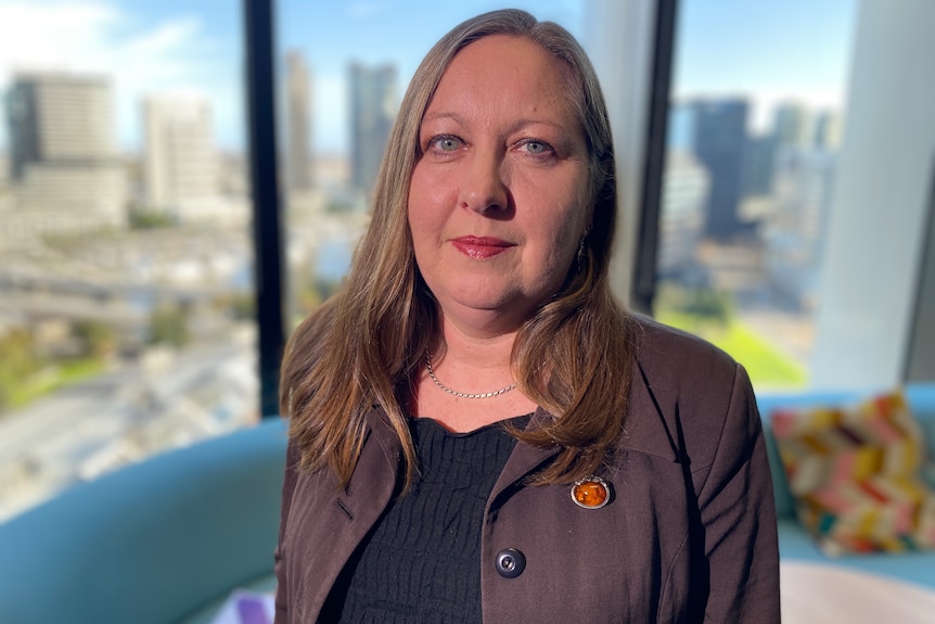 A middle aged woman with shuolder length mousy brown hair stands in an office with a sunny CBD in the background.