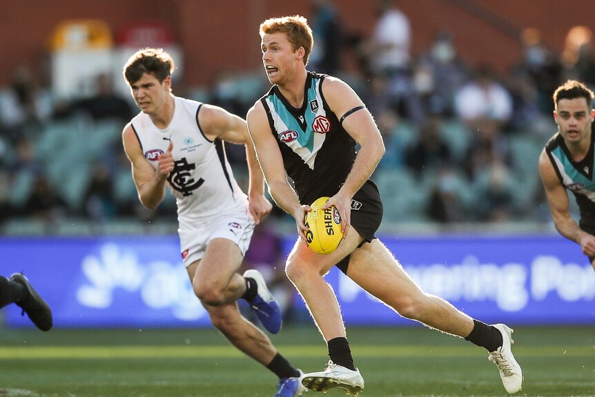A Port Adelaide player runs with the ball as a Carlton player chases behind him.
