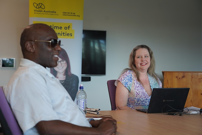 Sarah Evans sitting at a meeting table, smiling at a man wearing sunglasses.