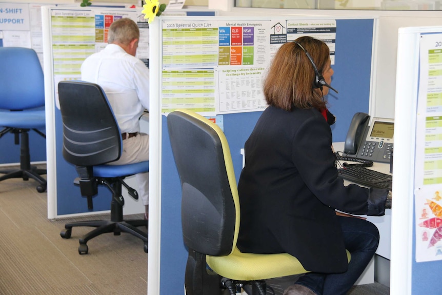 A woman and a man are each seated in a cubicle. The woman is wearing a phone headset.