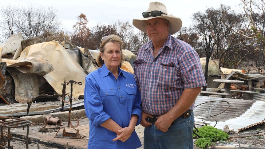 A woman in a blue shirt and a man in a checked shirt wearing a hat stand in front of what remains of a burnt house