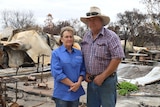 A woman in a blue shirt and a man in a checked shirt wearing a hat stand in front of what remains of a burnt house