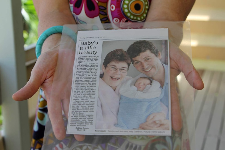Ange Newton holding a newspaper article about a baby born to parents who met at Beaut Blokes.