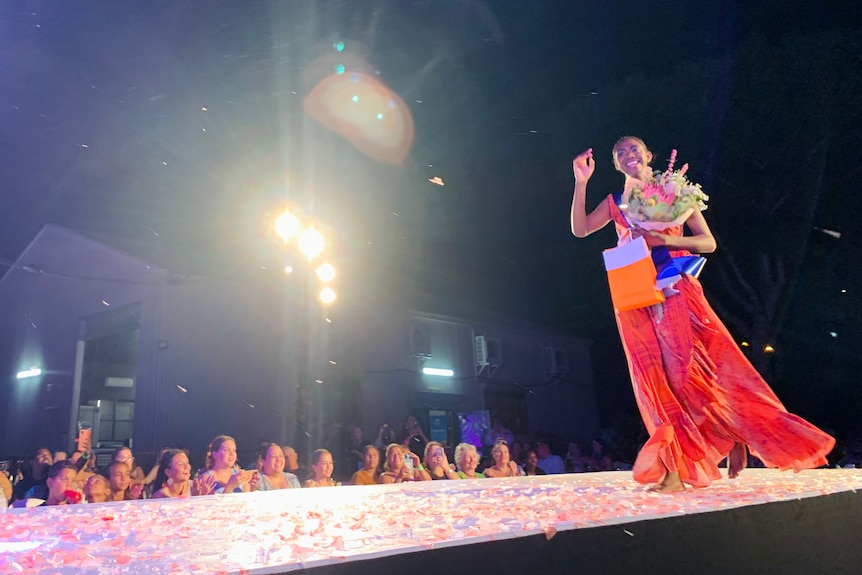 A wide shot of a catwalk stage with a woman in a dress waving and smiling as she walks.