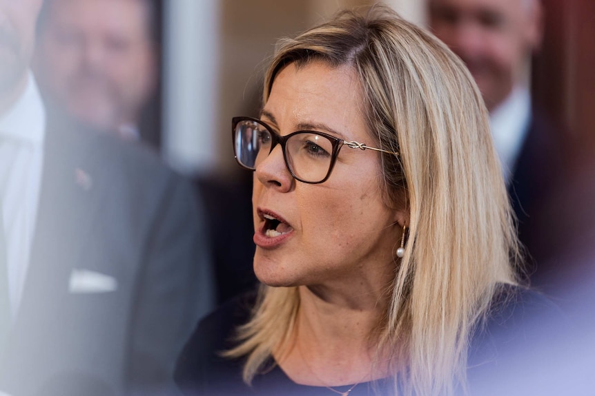 Close up of a blonde woman with glasses speaking to media