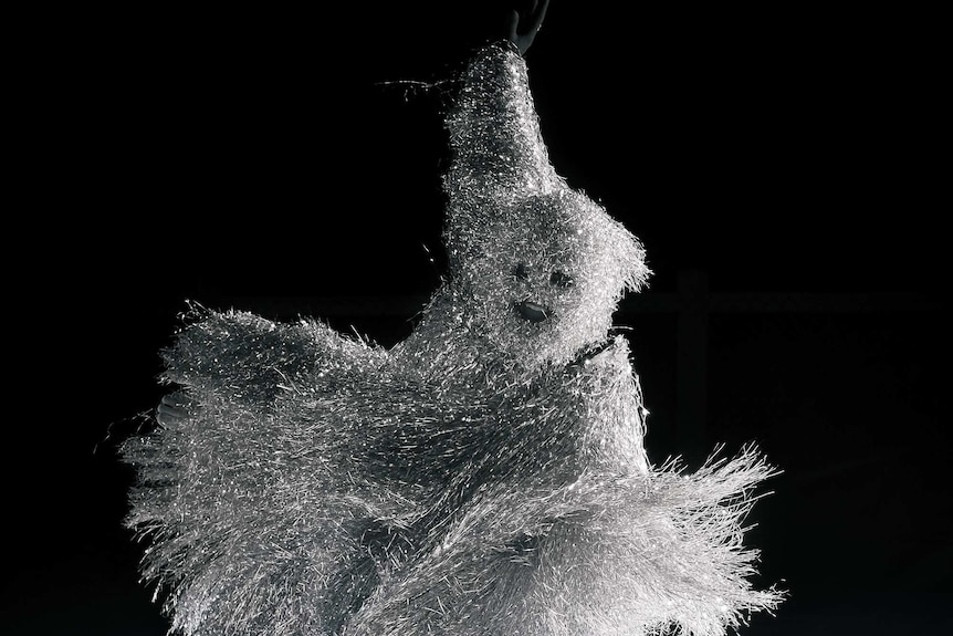 A person dressed in silver tinsel dances on white sand.