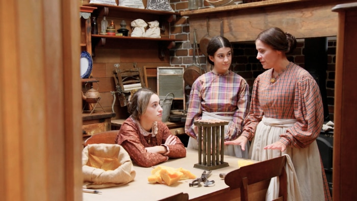 A photo of a mother and her two daughters dressed in old fashioned clothing in the back room of a corner store.