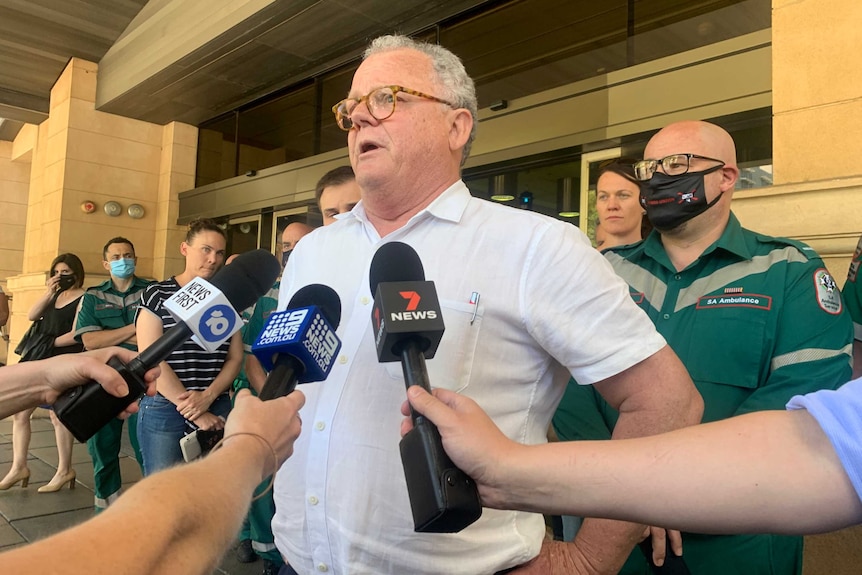 A man in a short-sleeved collared shirt and round glasses addresses reporters outside of court.