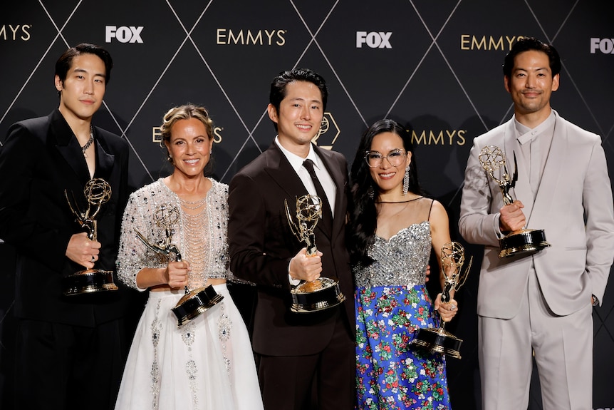 Five people stand in front of a black background each holding gold awards.