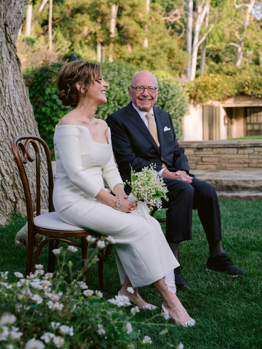 Elena Zhukova is seen smiling in profile, with Rupert Murdoch in background as both sit on chairs in a garden in formal dress