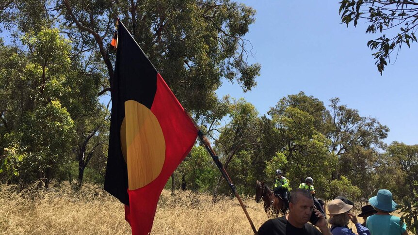 A protester flies the Aboriginal flag in bushland at Beeliar wetlands, with mounted police in the background.