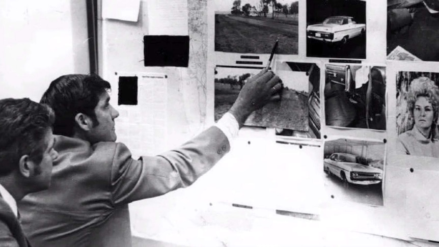 A black and white photo showing the back of two men pointing at an investigation pinboard