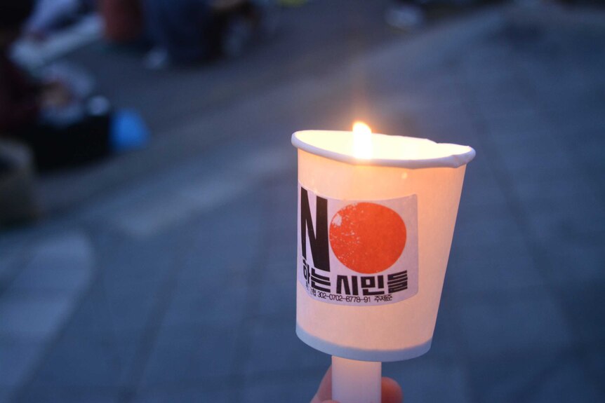 A close up of a candle flame sticking out of a cup reading: 'No Japan' in Korean.